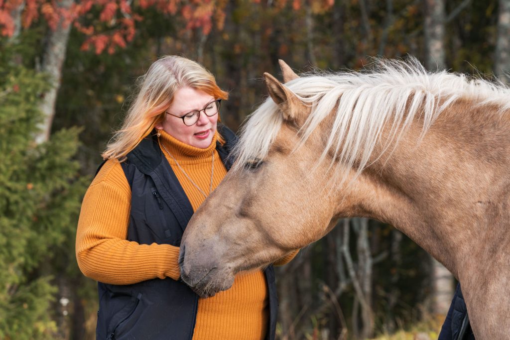 Sanna Kallunki seisoo vaalean hevosen vierellä syksyisessä ympäristössä.