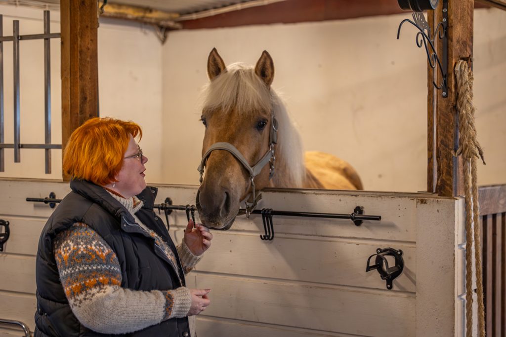 Sanna Kallunki seisoo vaalean hevosen vierellä tallissa.